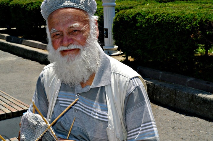 A smiling man with white beard sitting on a bench holding knitwear.