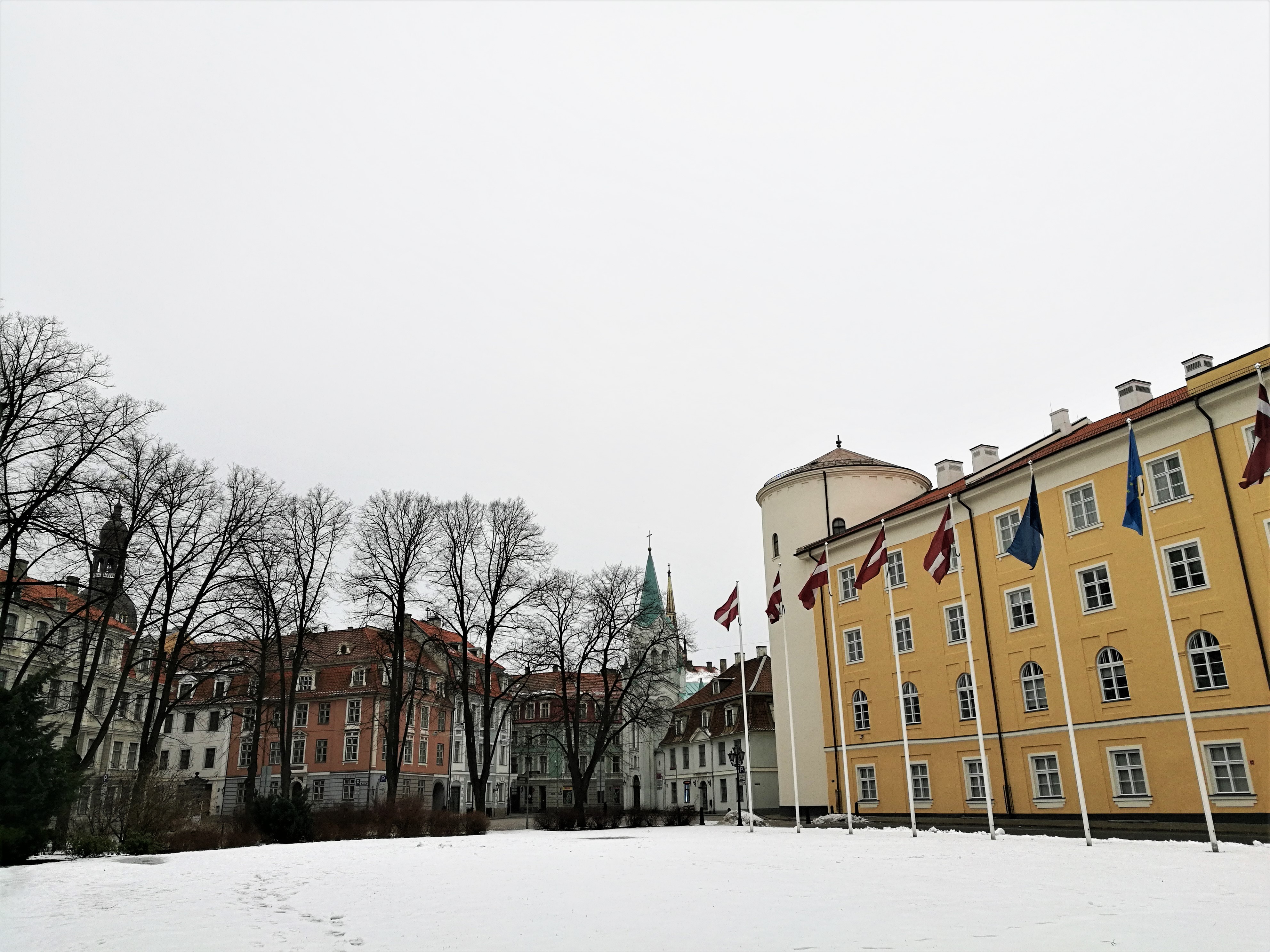 The yellow Riga Castle with a row of flags in front to the right, park with snow in the middle, and to the left, partly covered by leafless trees apartment buildings in pastel colours.