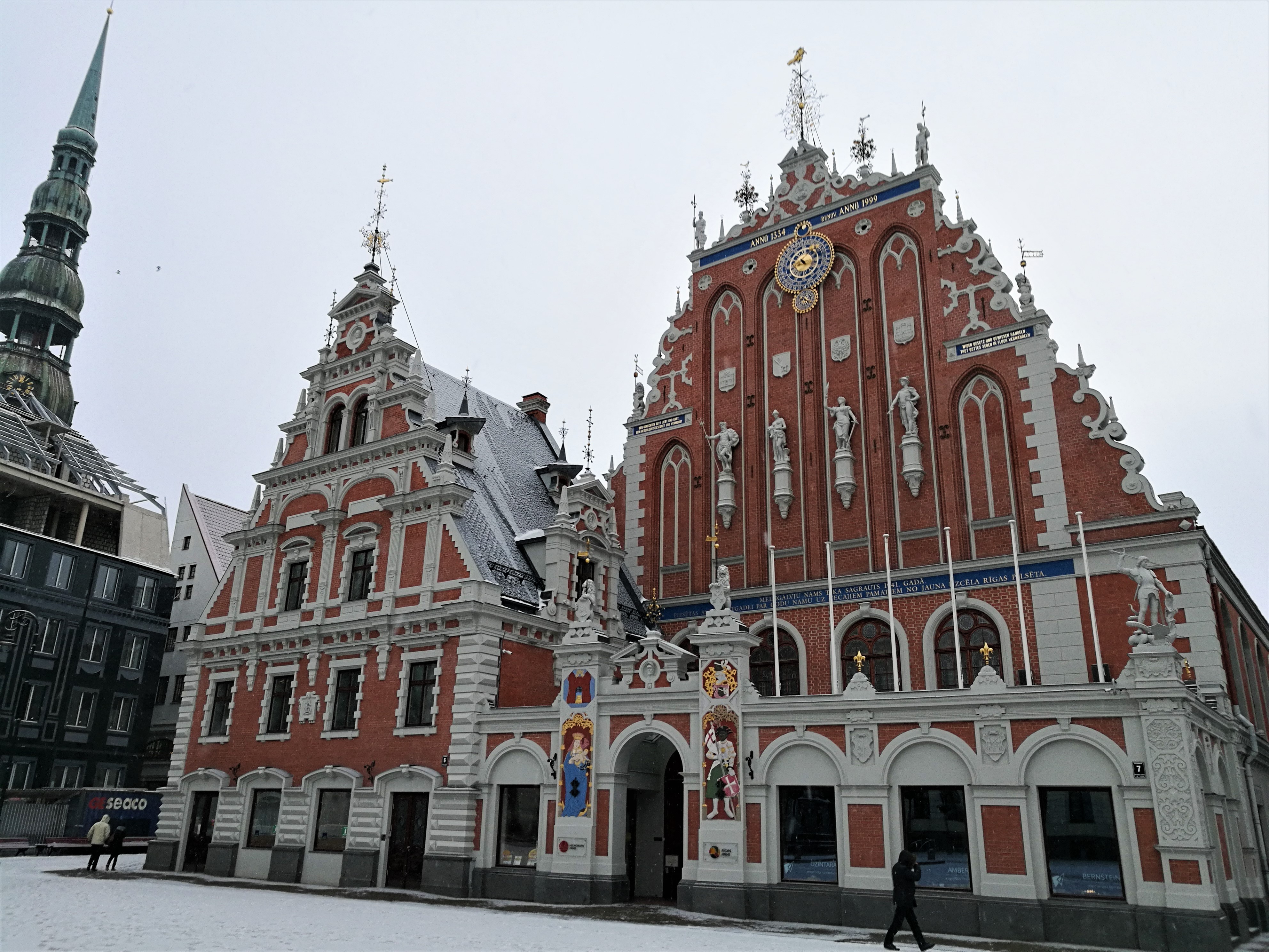 Picture of the House of Blackheads in Riga. Snow on the ground in front and a few people walking towards the sides of the building.