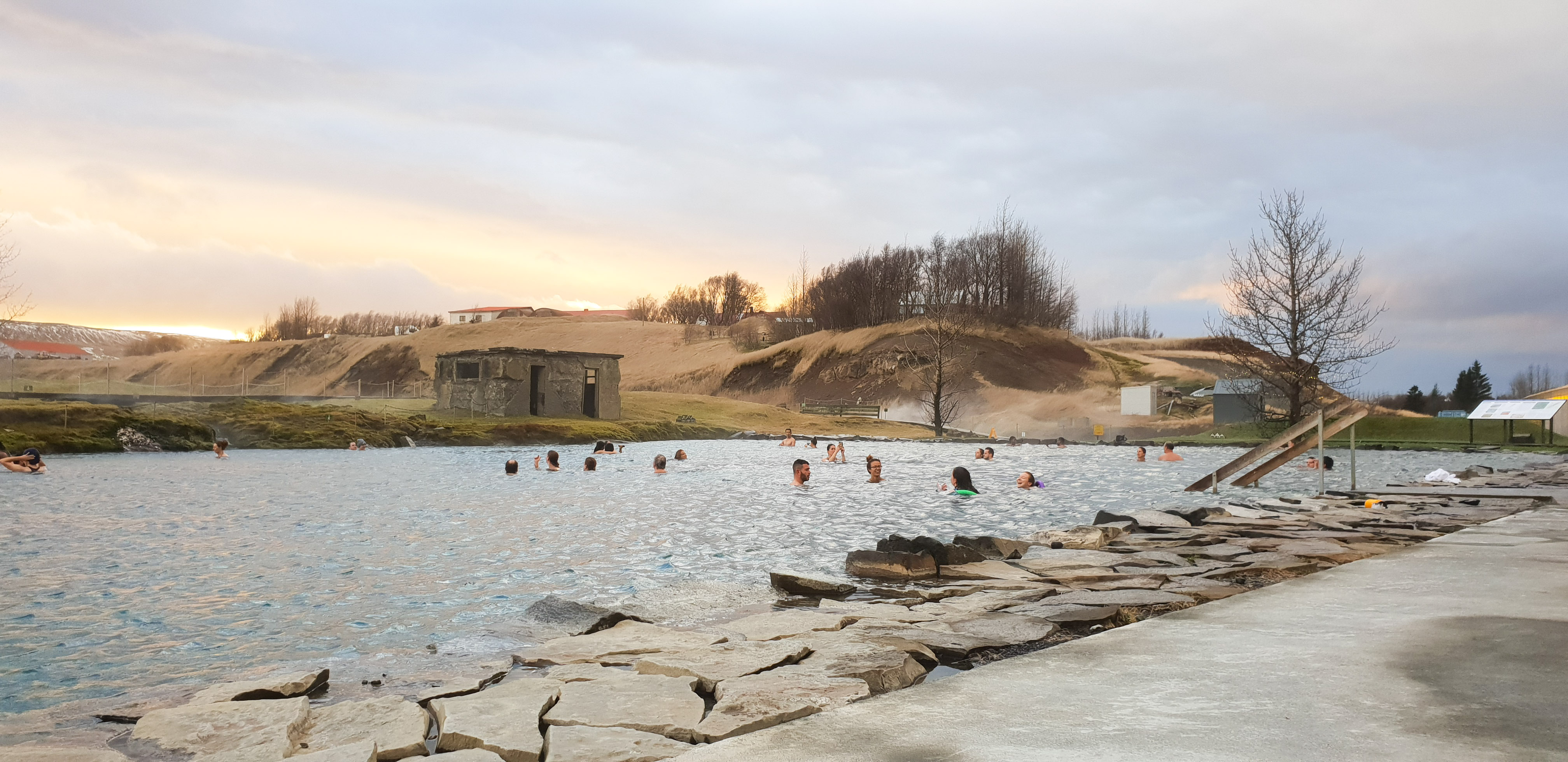 People bathing in the Secret Lagoon with a small hill and sunset in the back.