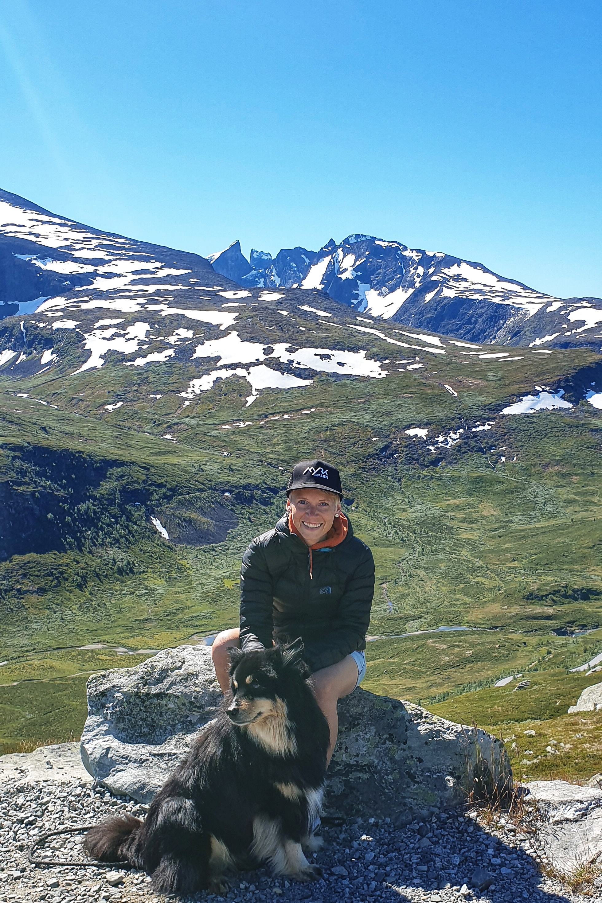 A woman in down jacket and jeans shorts with a dog in front of Skagastøltindan on Sognefjellet Scenic Route
