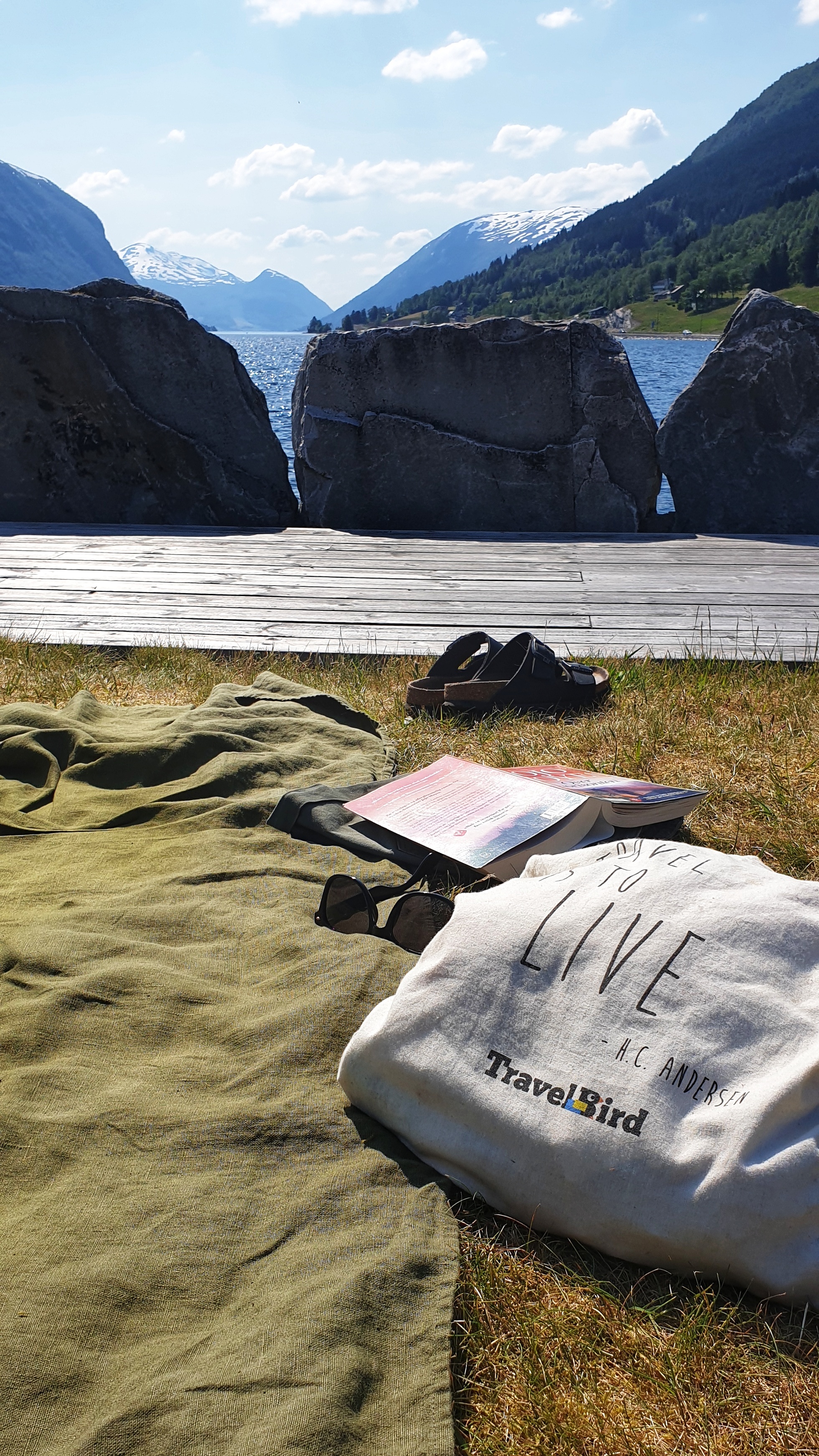 A cotton bag, a linen towel, book, sunglasses and sandals on a quay looking out on JØlstravatnet lake