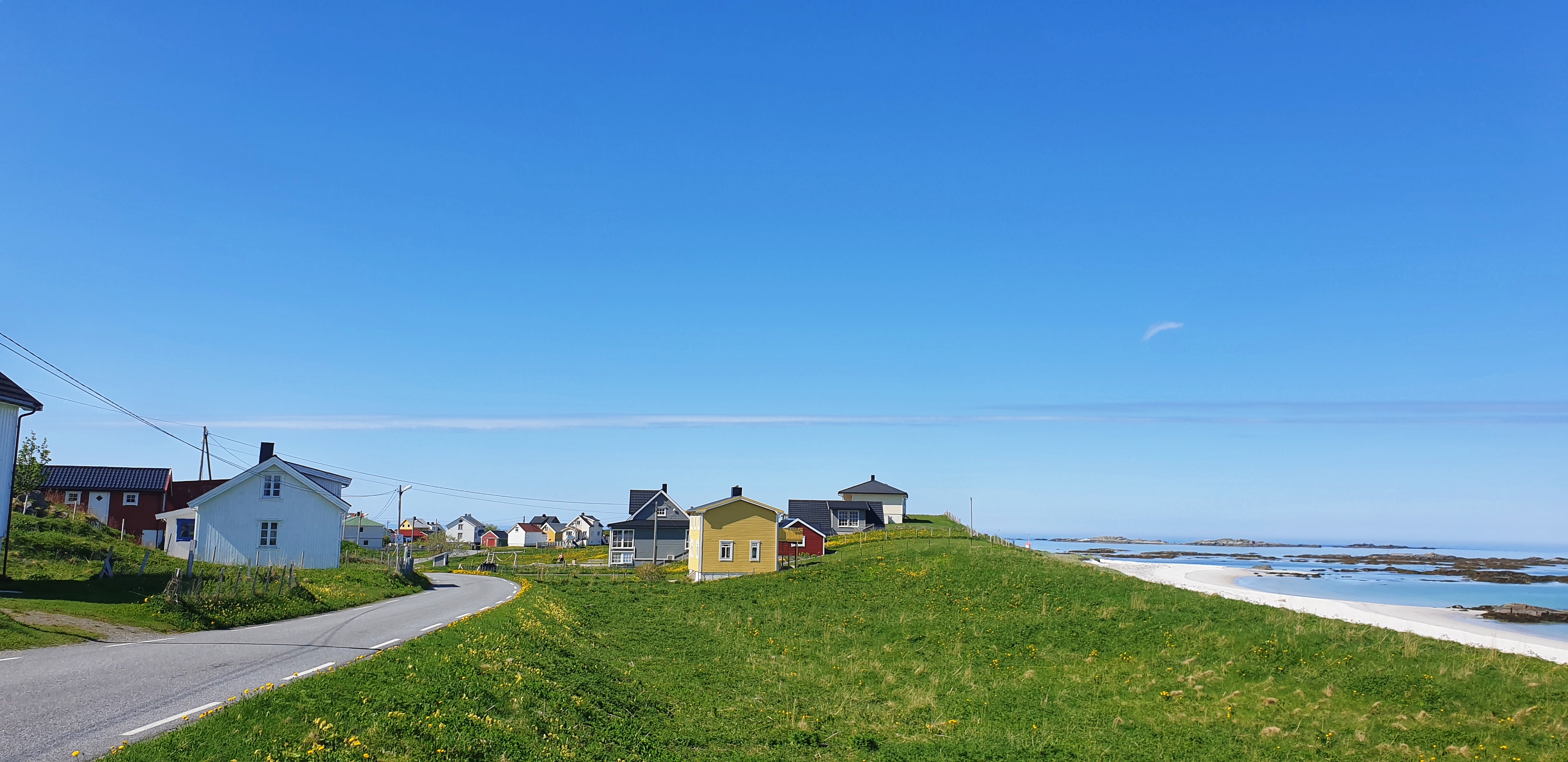 A road going between colourful houses next to green field, white beach and sea.