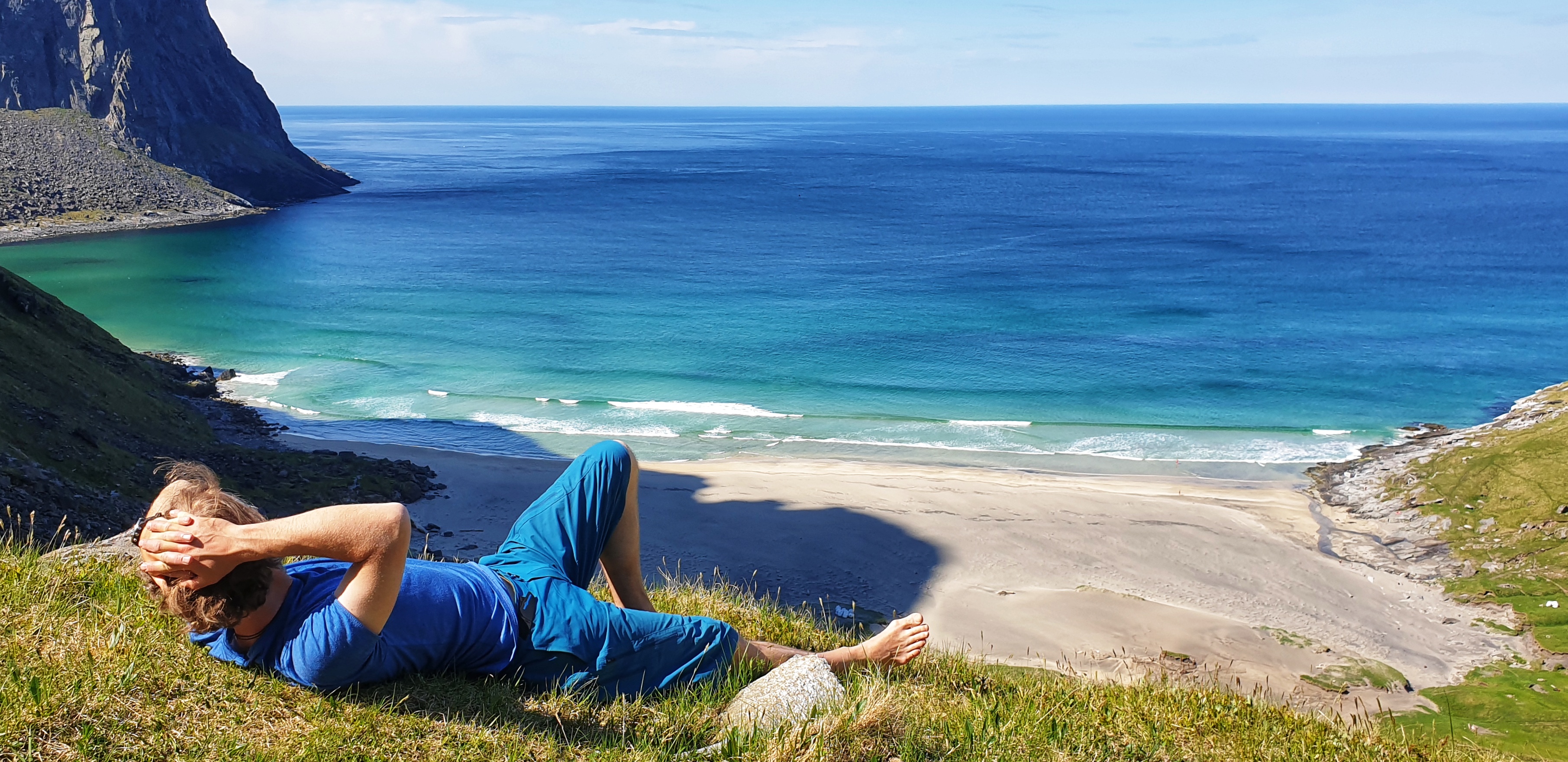 A man blue t-shirt and shorts lying on grass over Kvalvika beach and blue sea.