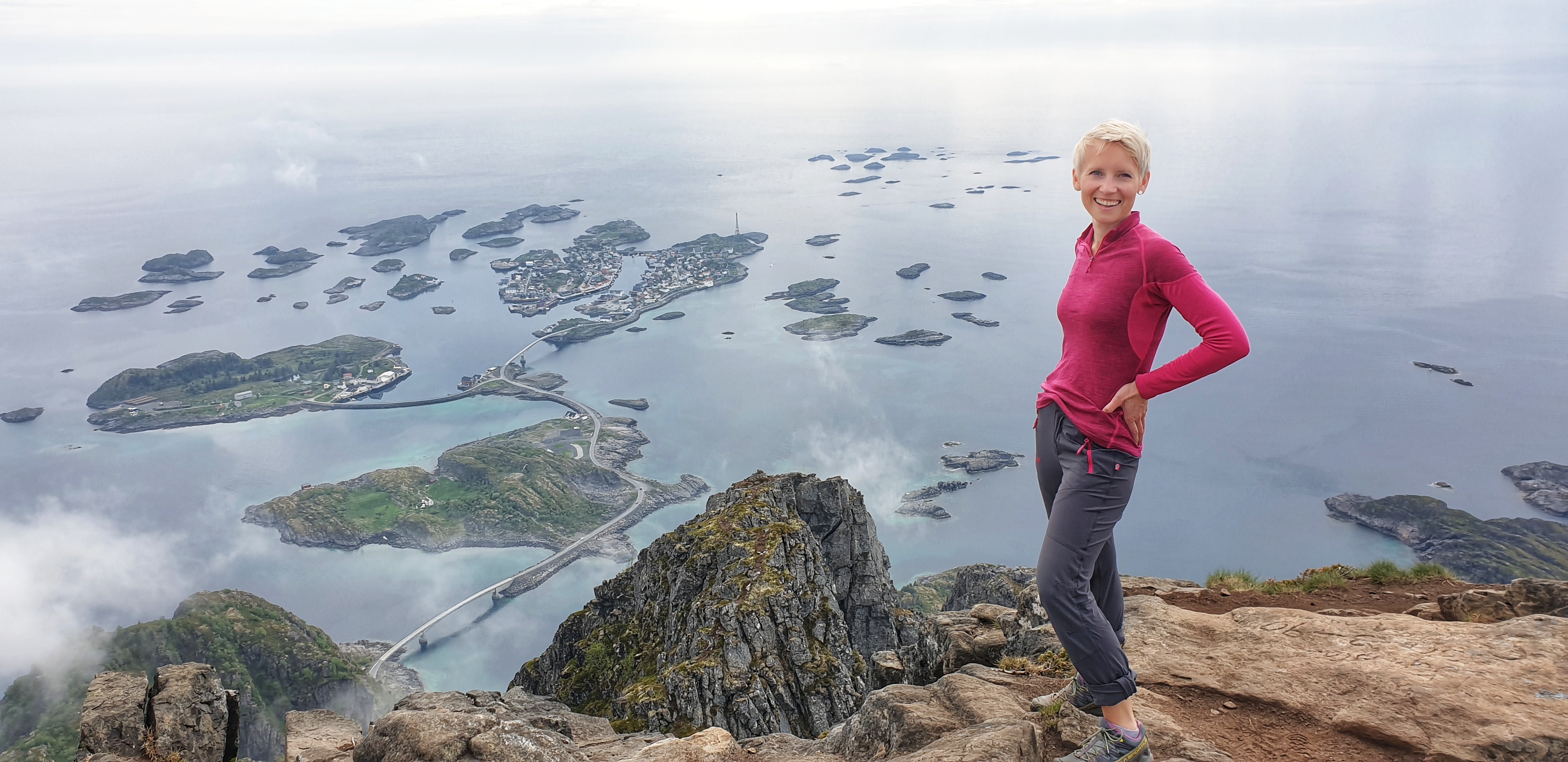 A woman in hiking outfit standing on a mountain. The sea and HenningsvÆr beneath.