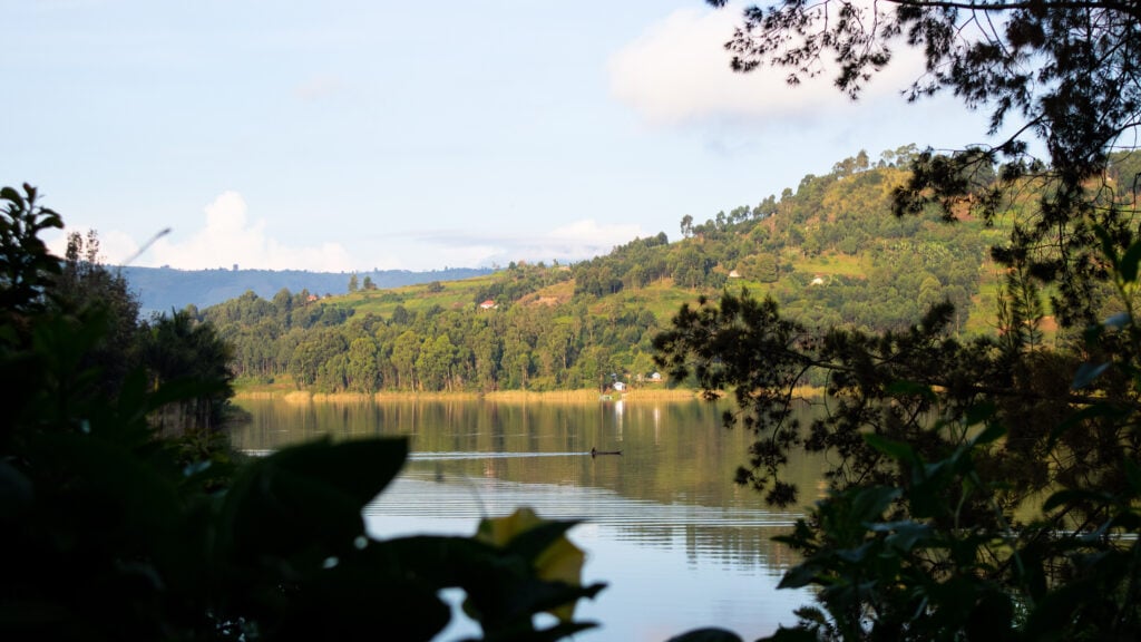 A canoe seen on Lake Bunyonyi through a frame of leaves. Forest, green hills and a few houses seen in the back.