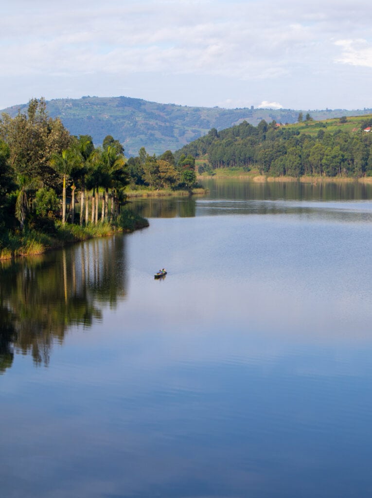 The view from a balcony on a day in Lake Bunyonyi. Canoe with two people, palm trees, forest, hills.