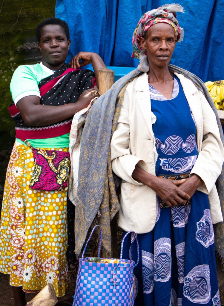 Two Ugandan women in colourful clothing in front of a stand with bananas looking into the camera.