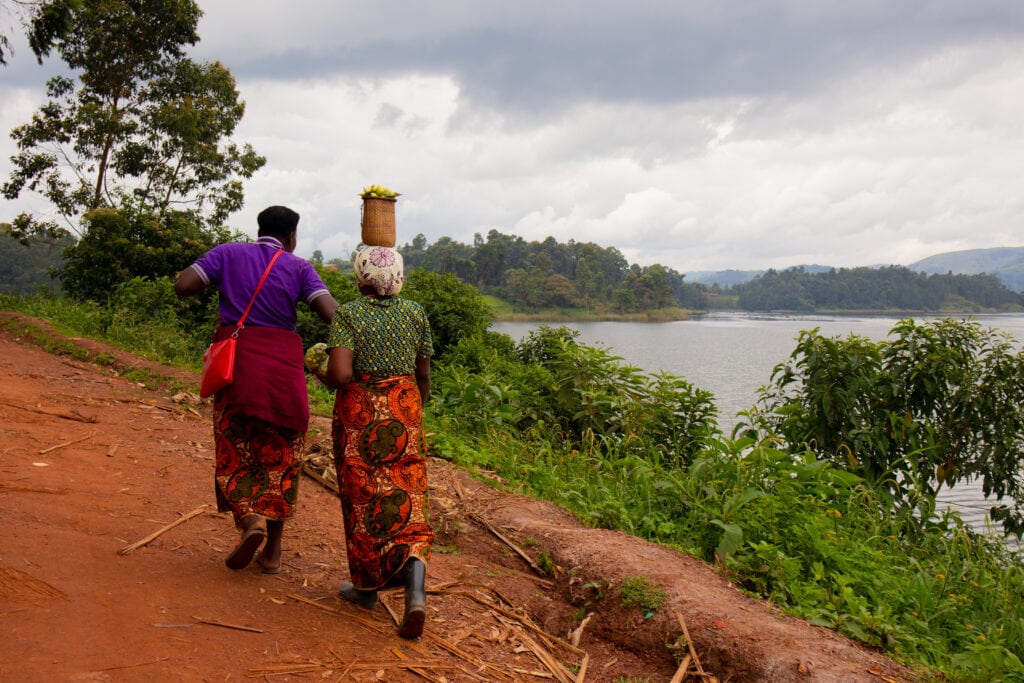 Two women in colourful clothes, one carrying a basket on her head, on a dirt road by Lake Bunyonyi.