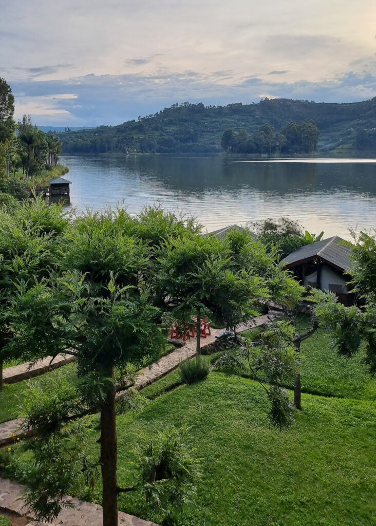View of the garden of Bunyonyi Overland Resort and some roofed tents. Lake Bunyonyi and green hills seen in the back.