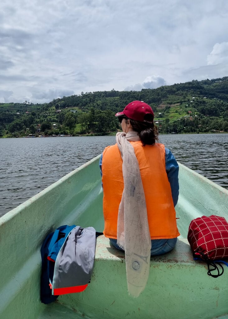 A woman wearing a life west, scarf, and red hat sen from behind sitting in a boat on a day trip on Lake Bunyonyi.