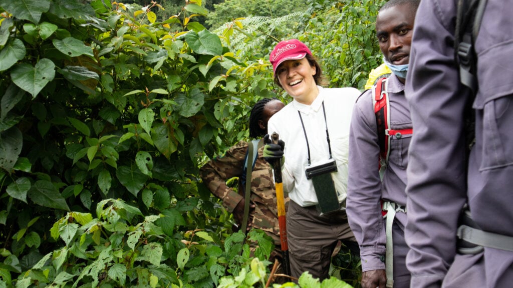 A woman in red hat and white bluse smiling at the camera, holding a walking stick. A man also looking at the camera. A male body spotted in front and a woman behind. Dense bushes around.