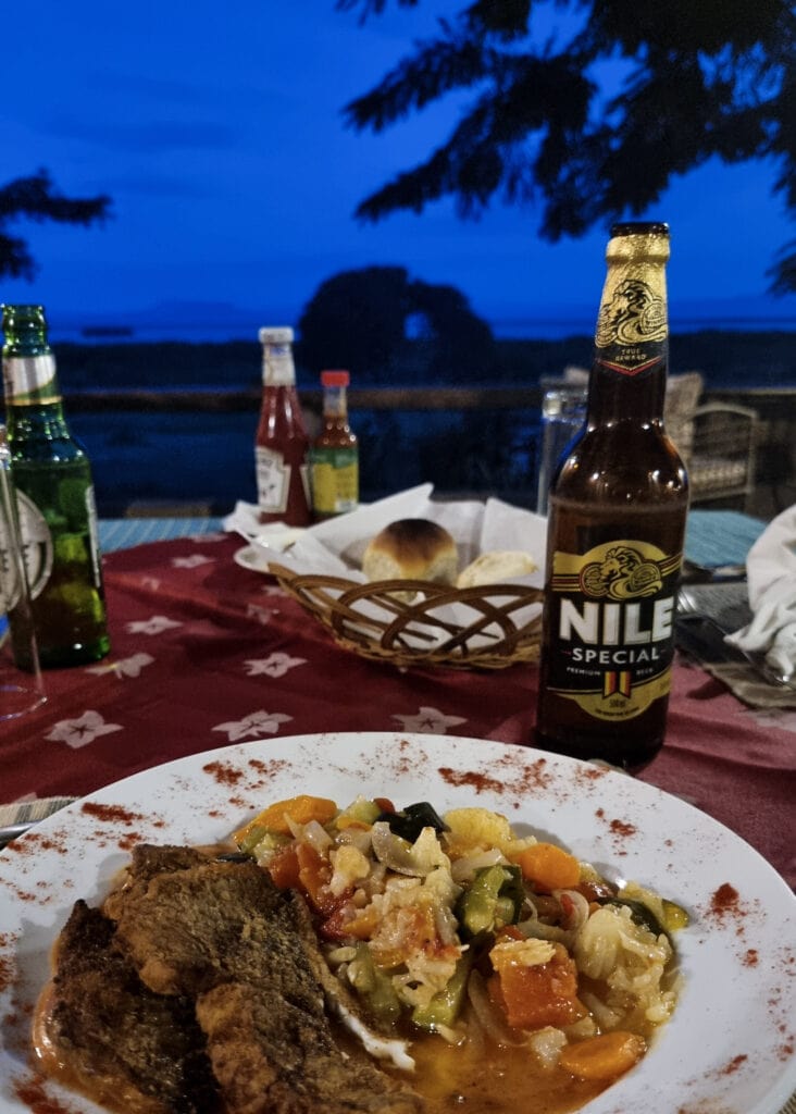 A plate with food, two beer, and a bread basket on a table. Views towards Lake Edward in dusk.