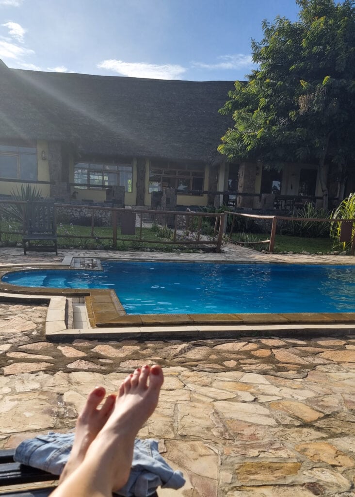 To feet on a sun lounge chair in front of the swimming pool and main building of Ihamba Safari Lakeside Lodge