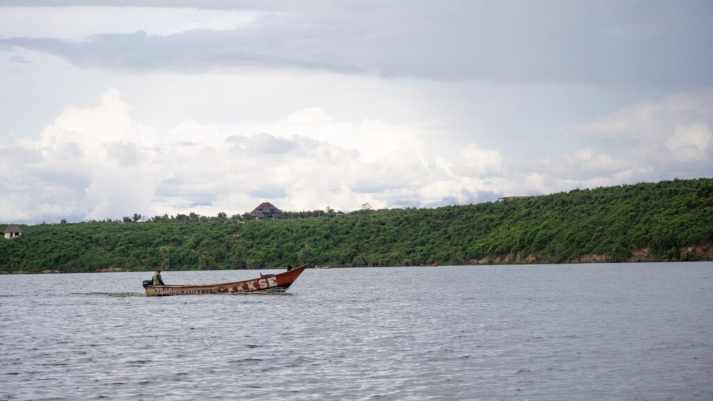A red wooden motorboat with white writing on the side with two men on board on the Kazinga Channel. Blue sky, green forest and a large building behind.