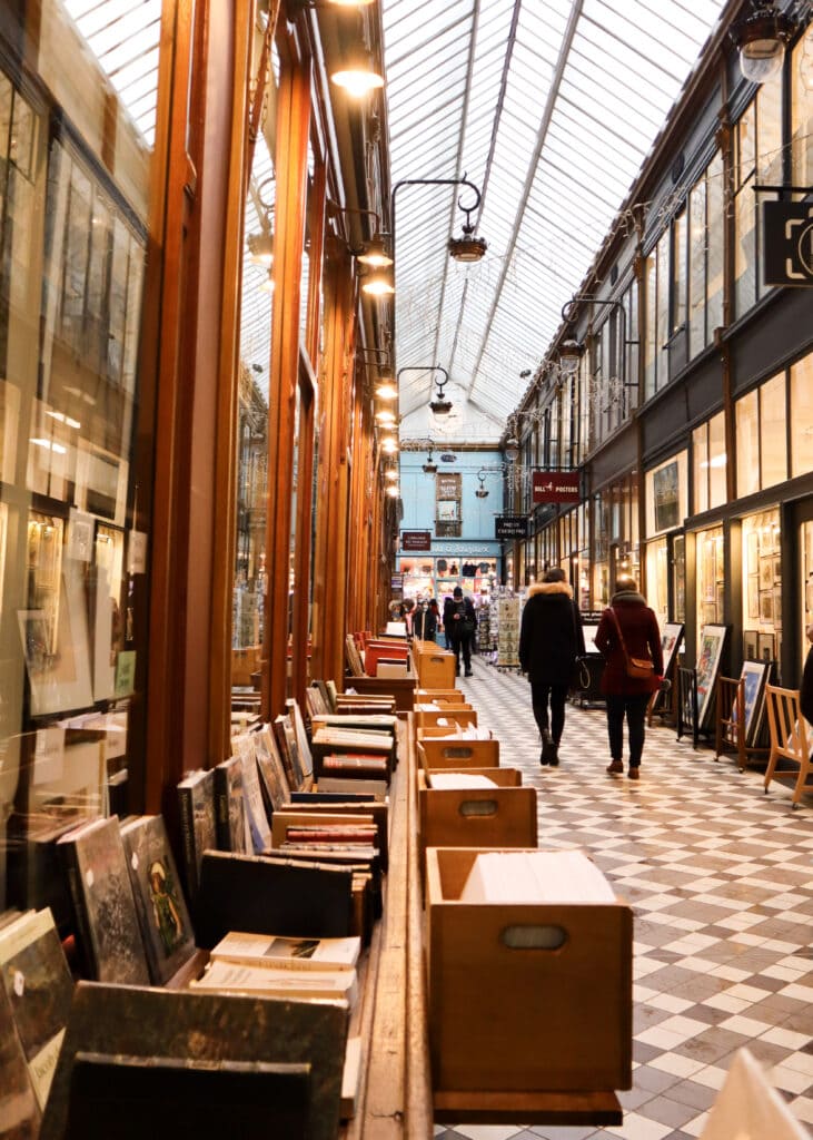 People walking inside Passage des Panoramas. Books on a shelf and in boxes in the front.
