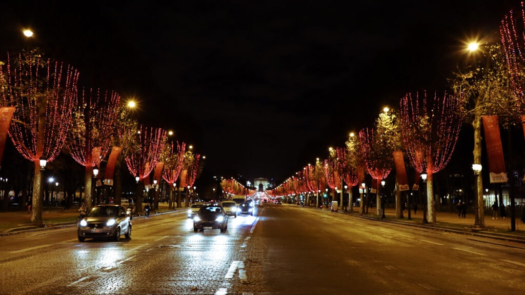 Cars driving on Avenue des Champs-Élysées, red lights covering the trees on both sides. Arc the Triomphe in the back.