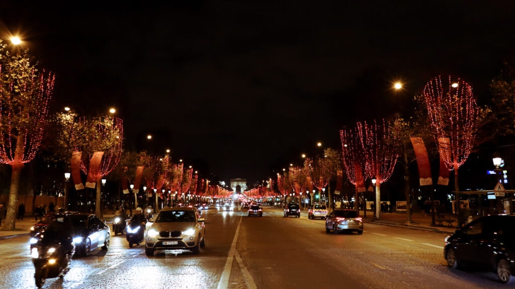 Cars driving on Avenue des Champs-Élysées, red lights covering the trees on both sides. Arc the Triomphe in the back.