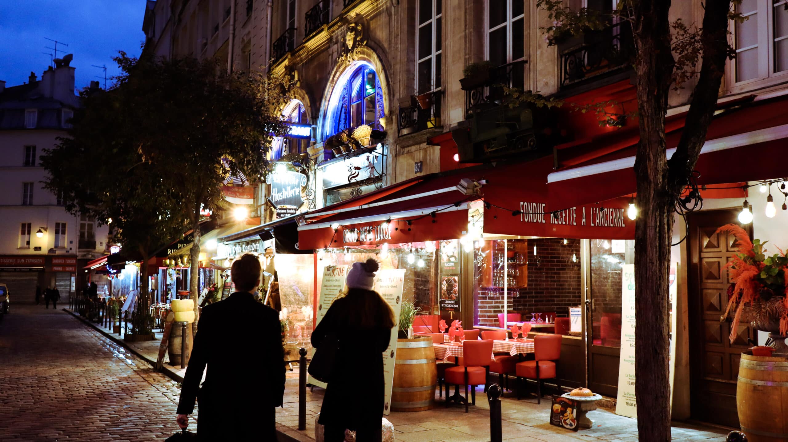 Two people walking on a street in Paris. Restaurants and colourful lightning around.