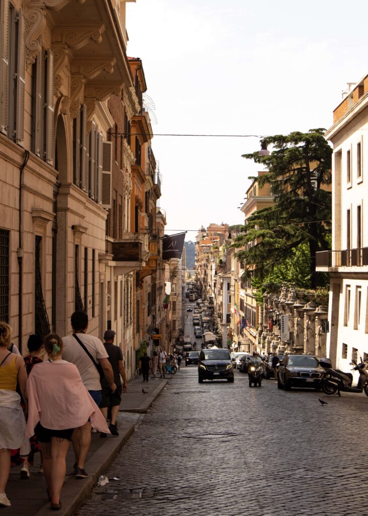 People walking on a sidewalk along a street in Rome in dim sunlight.
