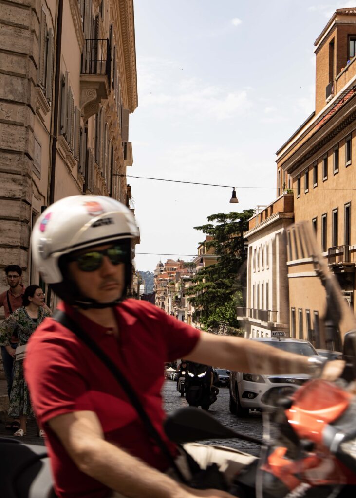 A man in red t-shirt and white helmet driving a scooter in Rome.