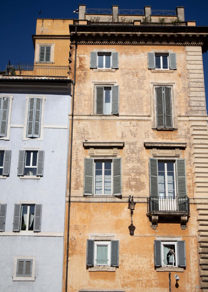 Two pastel coloured buildings with window shutters in morning light in Rome. A man can be seen on the lower right window.