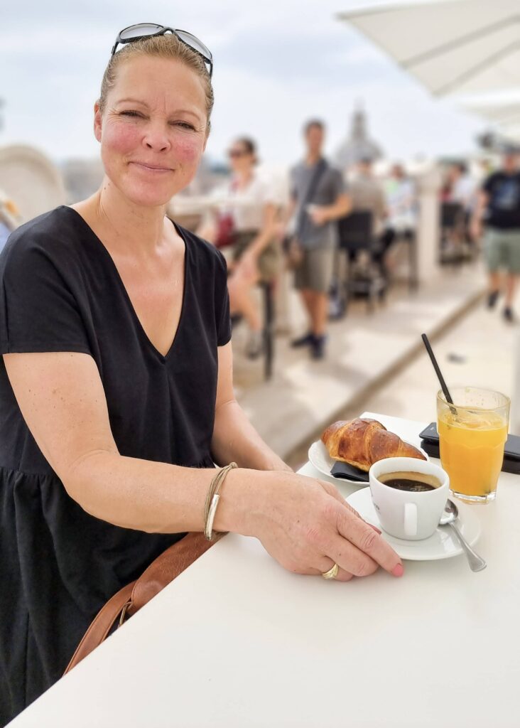 A woman in black dress smiling at the camera, a cup of coffee, glass of orange juice and a croissant on a table.