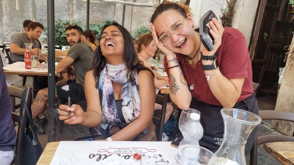 Two women laughing by a restaurant table.