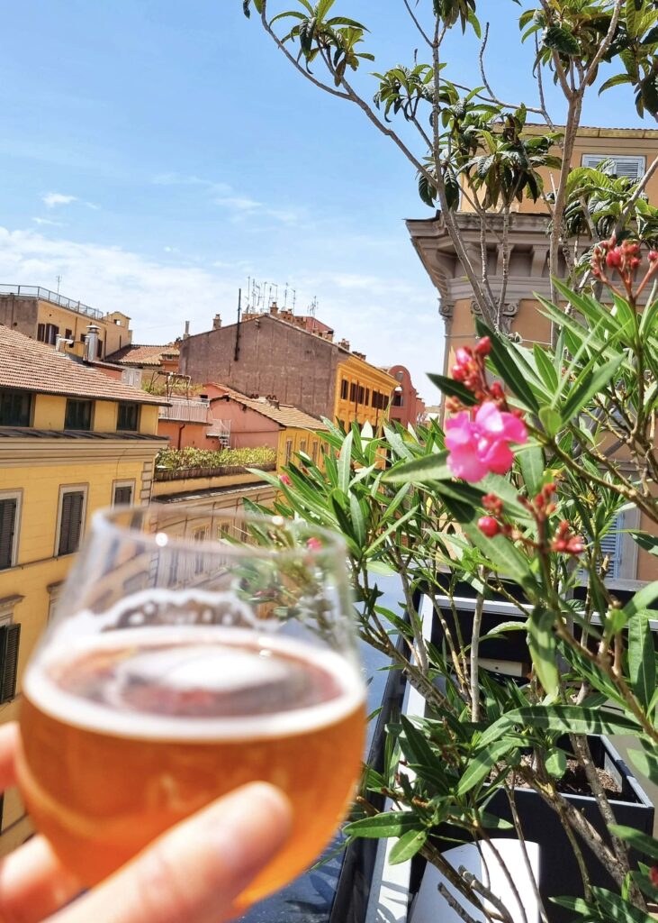 A glass of beer in front of pink flowers and colourful buildings in Rome.