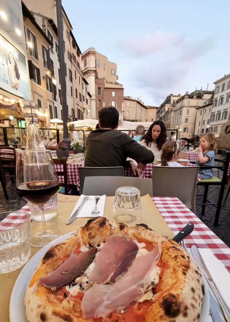 A plate with pizza and a glass of red wine in front of views of Campi de Fiori.
