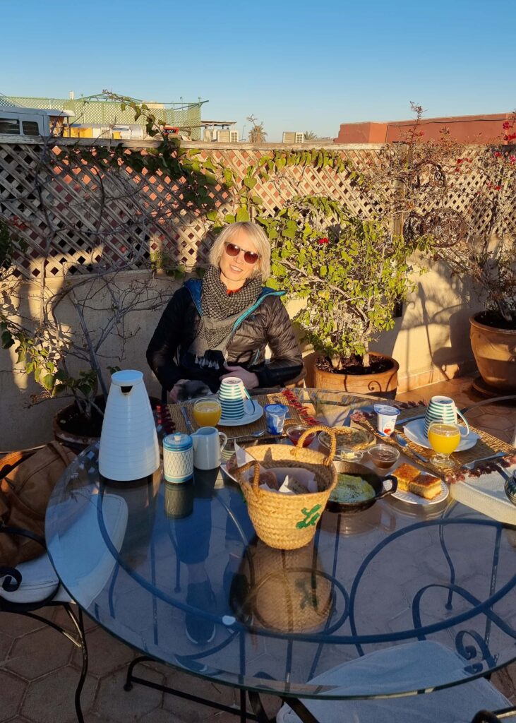 A woman wearing down jacket, skarf, and sun glasses sitting behind a glass table filled with a Moroccan breakfast.