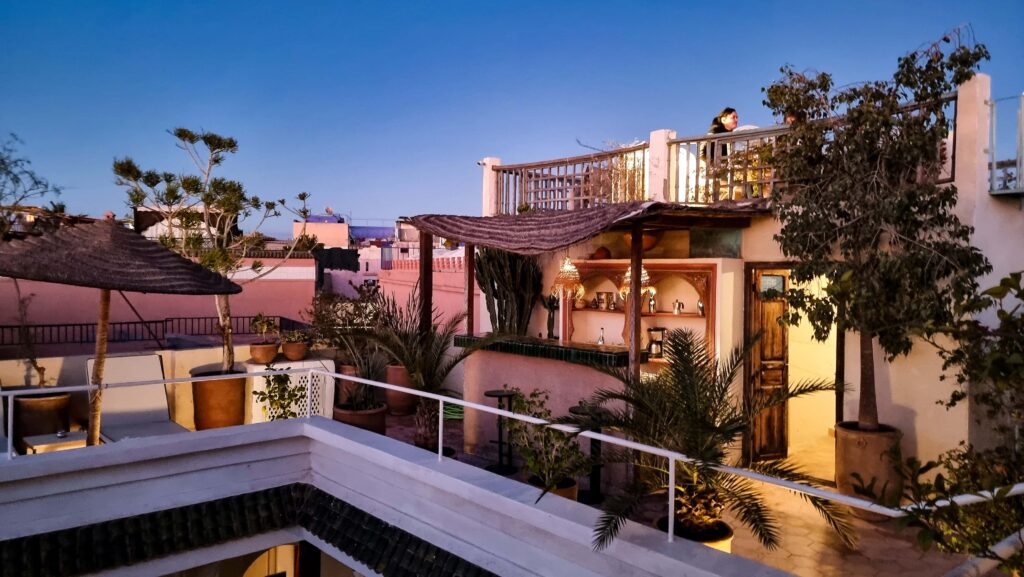 Riad Le Coq Berber Fou's rooftop terrace at dusk. A bar, palms and other plants and sun loungers can be seen.