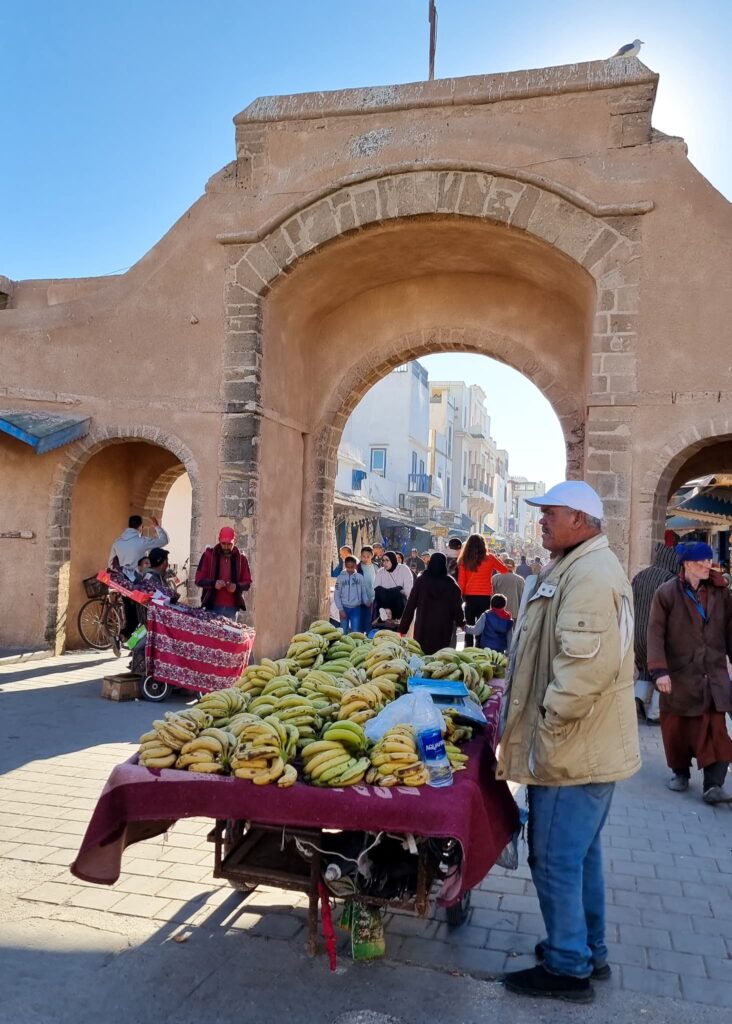 Street life in Essaouira during 1 weekn in Morocco. A banana seller in front, people walking in and out of a gate behind.
