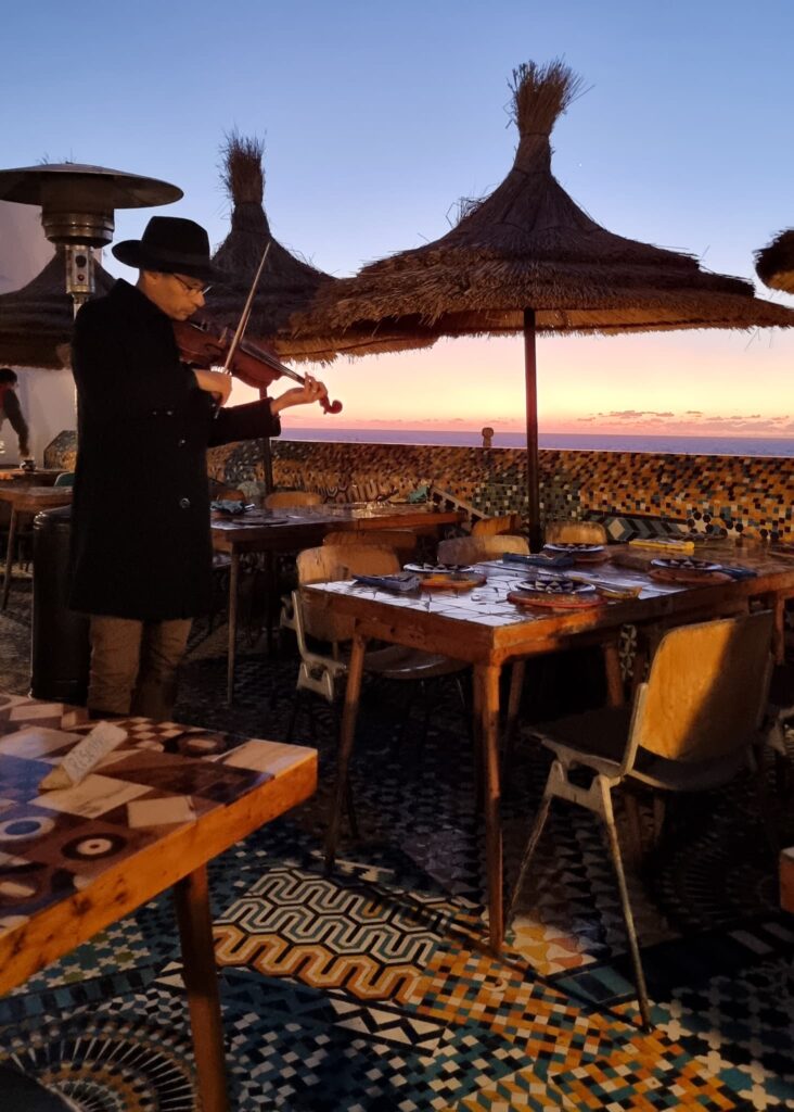 A man in black hat and coat playing violin amongst empty tables on Salut Maroc's rooftop terrace at sunset in Essaouira.