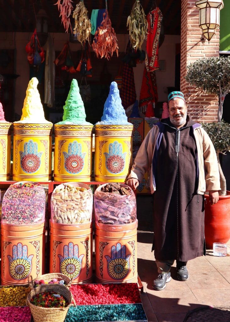 A man standing next to bins with coloured powder and herbs outside his shop in Marrakech.