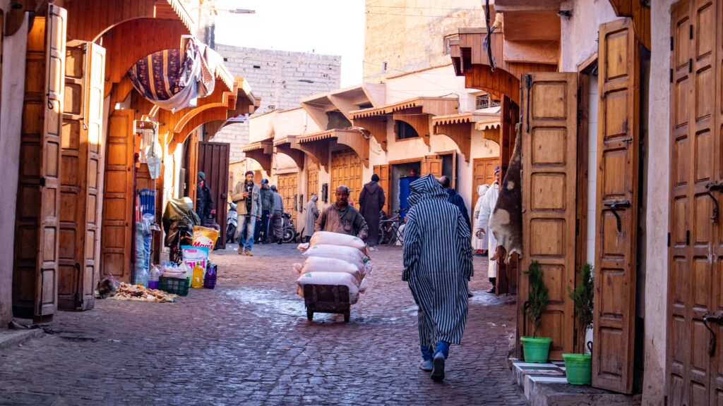 Street photo in Marrakech medina. A man in a striped djellaba seen walking from behind, a man with a trolley holding big white bags coming towards the camera, and people standing outside open wooden doors.