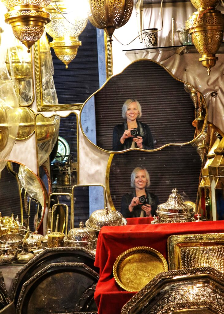 A shop in Marrakech souk with brass works. A woman holding a camera can be seen in two mirrors.