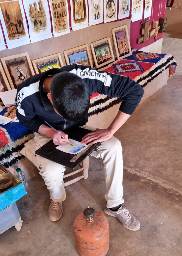 An artist painting with water colours. More paintings hanging on a wall behind him.