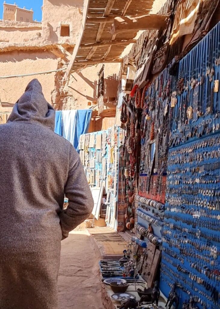 A man in a grey djellaba walking past jeelery hanging on a wall in Morocco.