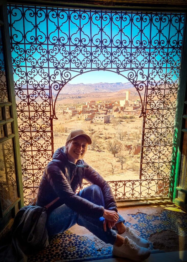 A woman sitting in a windowsill in Telouet Kasbah. A village with a mosque seen through a decorated window.