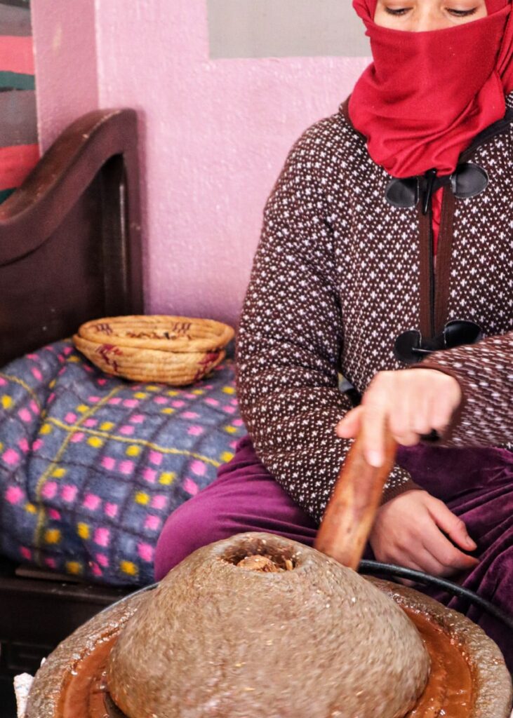 A woman wearing a red hijab and black and white knitted jacket extracting argan oil from nuts in the traditional way.
