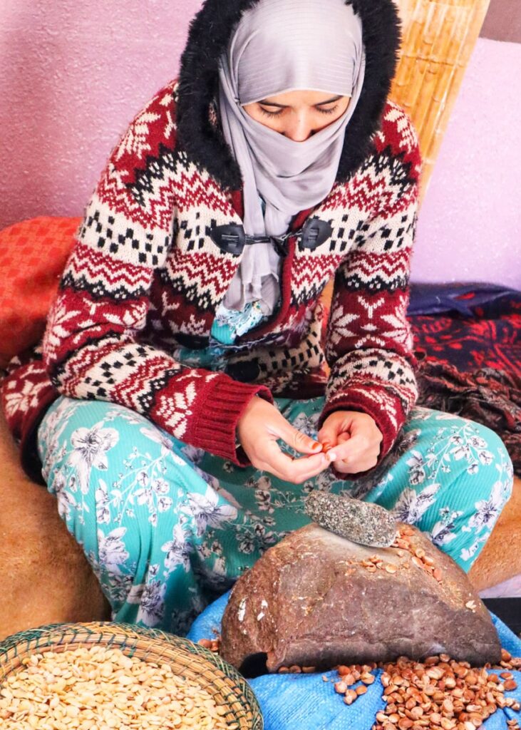 A young woman in a head scarf covering her nouse and a knitted cardigan pealing Argan nuts.