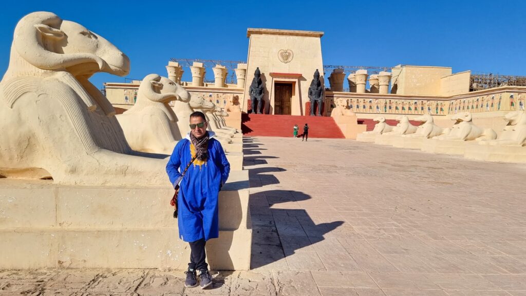 A guide wearing sunglasses and a blue and yellow shirt standing infront of a movie set from Asterix and Obelix in Ouarzazate's Atlas Studios.
