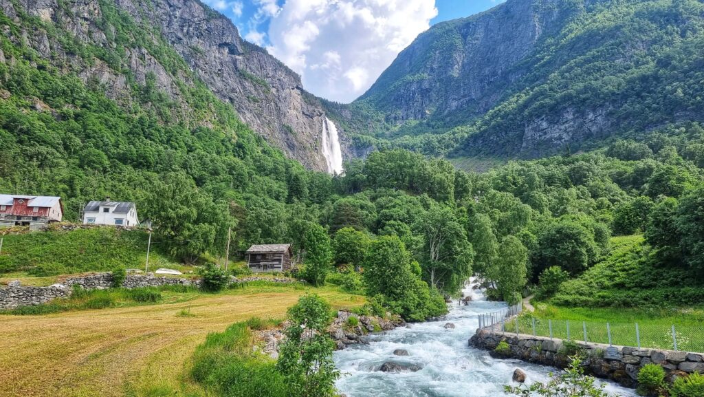 Feigefossen waterfall and a roaring, white river between large mounatins and green forest. A small farm to the left hand side.