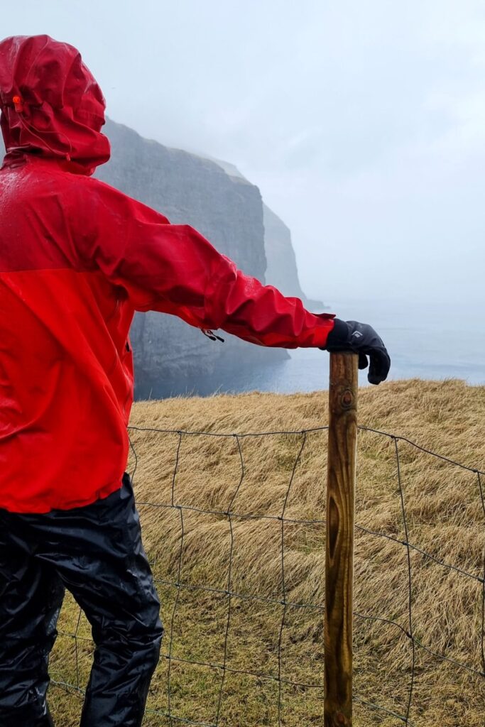 A man in dripping wet red rain coat and black rain pants standing by a fence looking towards the Faroe's coastline.