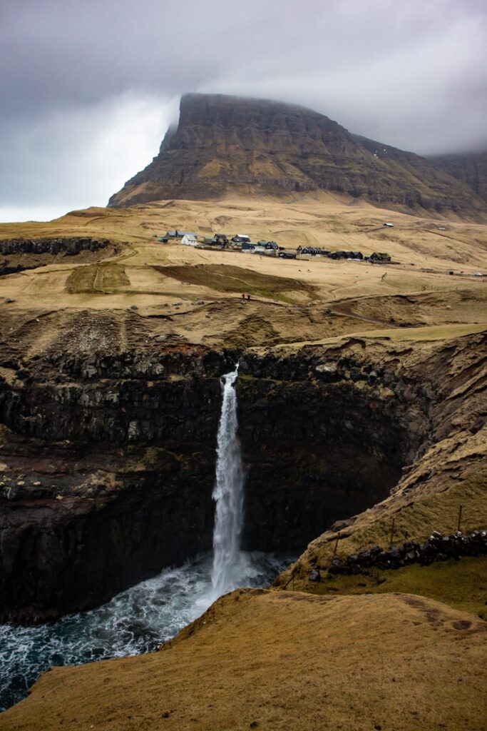 Waterfall plunging into the sea near a small village and a peak partly covered in fog on the Faroe Islands.