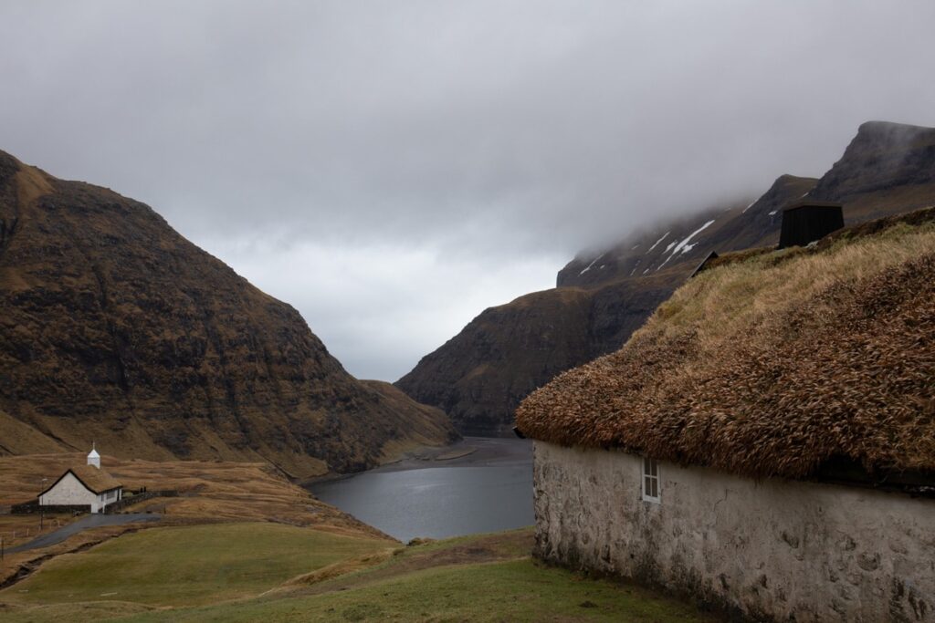 Old brick house with turf roof and a small church in Saksun, Bay and mounatisn in the background.