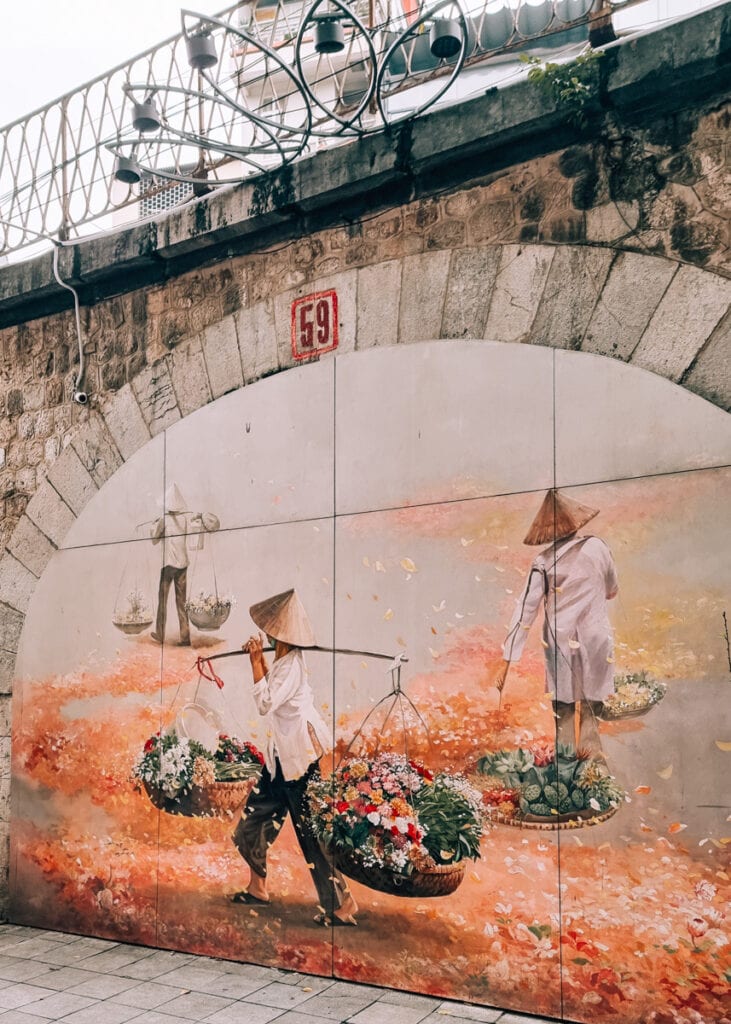 A street mural showing three traditional Vietnamese women wearing conical hats and carrying flowers and fruits from baskets hanging over their shoulders.