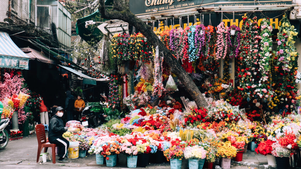 A flower shop in Hanoi busting with colourful flowres. A woman with face mask sitting on a chair next to it.