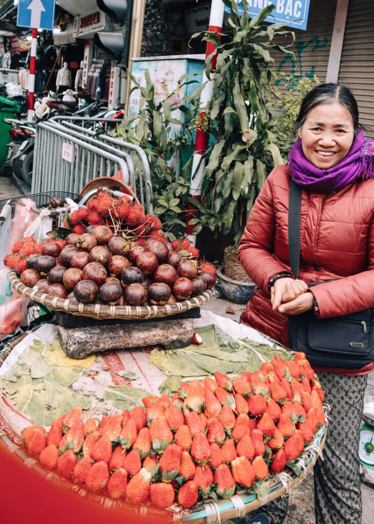 An older female street vendor in Hanoi, standing beside her bike filled with fruit and berries.